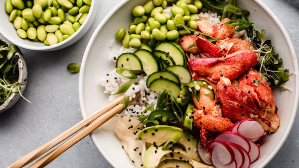 A lobster poke bowl with rice, sliced cucumbers, avocado, radishes, edamame, and fresh herbs.