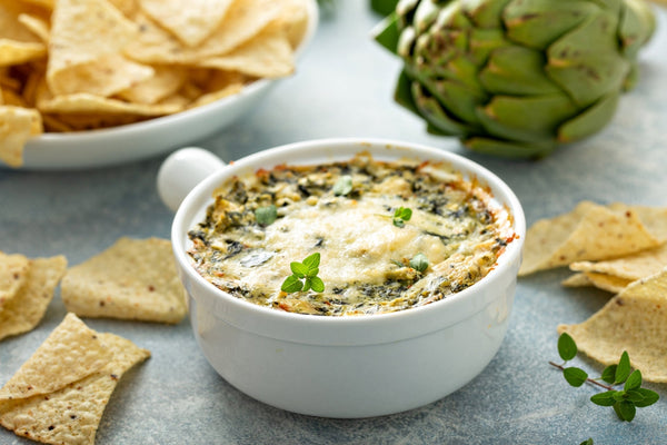 Warm, cheesy Maine crab and artichoke dip served in a white bowl with tortilla chips and a fresh artichoke.