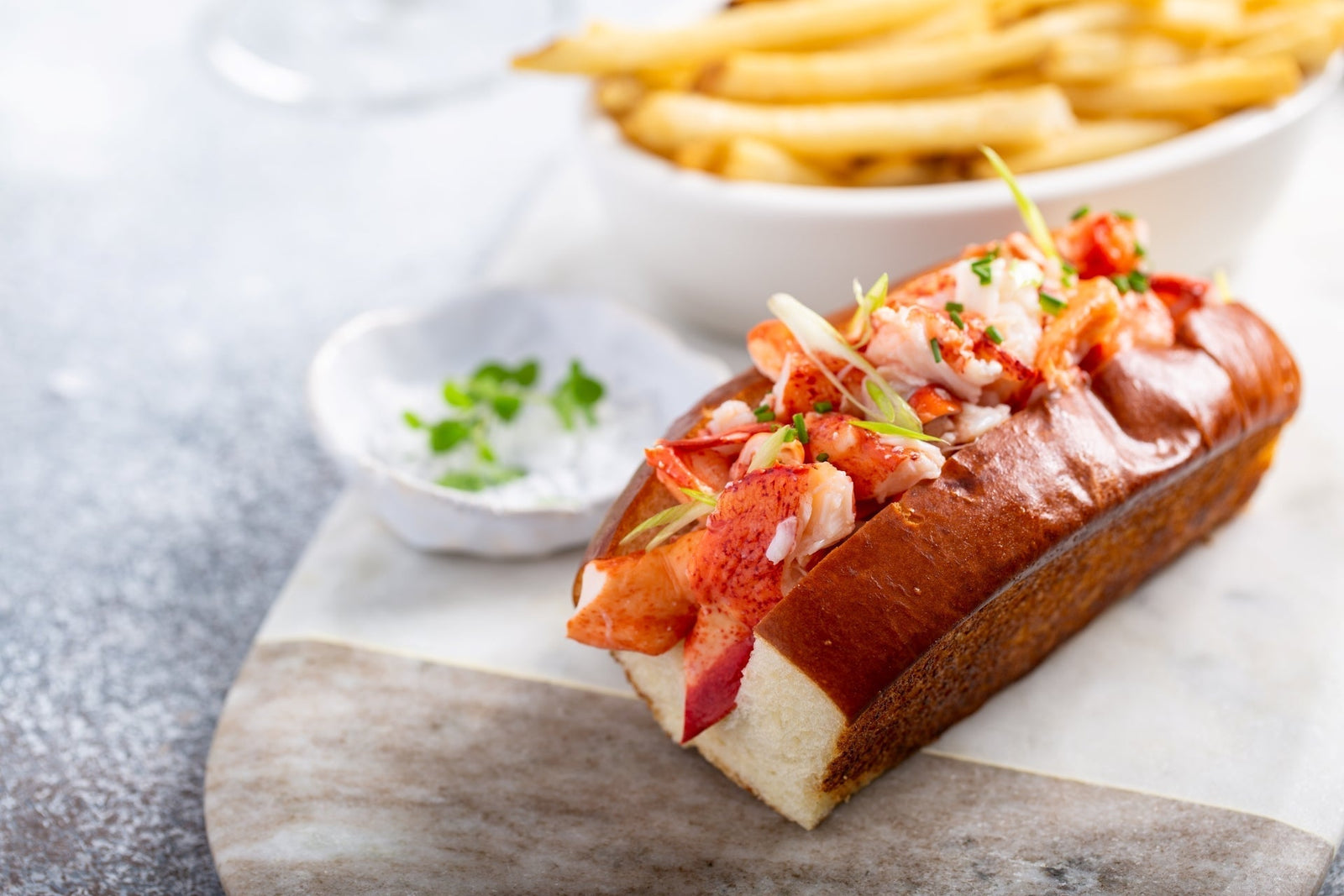 Split-top bun packed with fresh Maine lobster chunks, served with chives, dip, and crispy fries in the background.