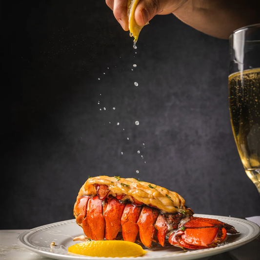 Sprinkling lemon on lobster tails, alongside a glass of champagne and a lemon wedge.