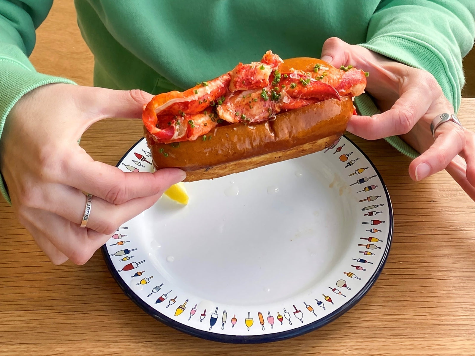 A person wearing a green sweater is holding a Chicago-style lobster roll while sitting near a wooden table.