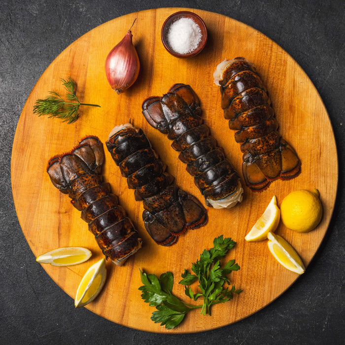 Four raw Maine lobster tails displayed on a wooden board with fresh parsley, dill, shallot, lemon wedges, ready for prep.