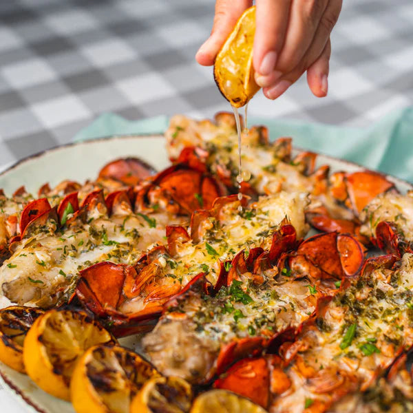 Platter of broiled lobster tails with herbs and lemon being drizzled on top.