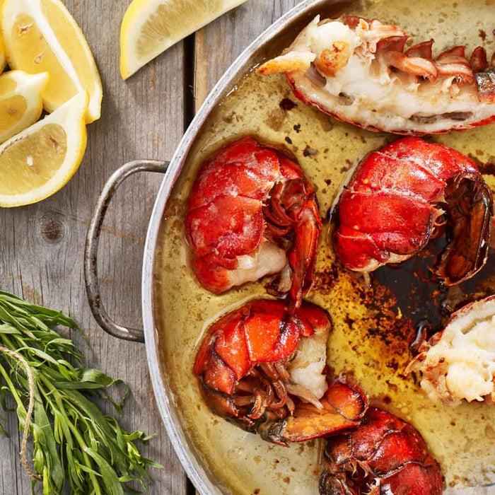 Grilled Maine lobster tails topped with herbs and butter, with lemon juice being squeezed over them for added flavor.
