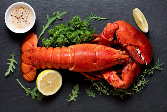 Cooked Maine lobster tail and claws served with lemon, dipping sauce, and fresh herbs on a black slate plate.