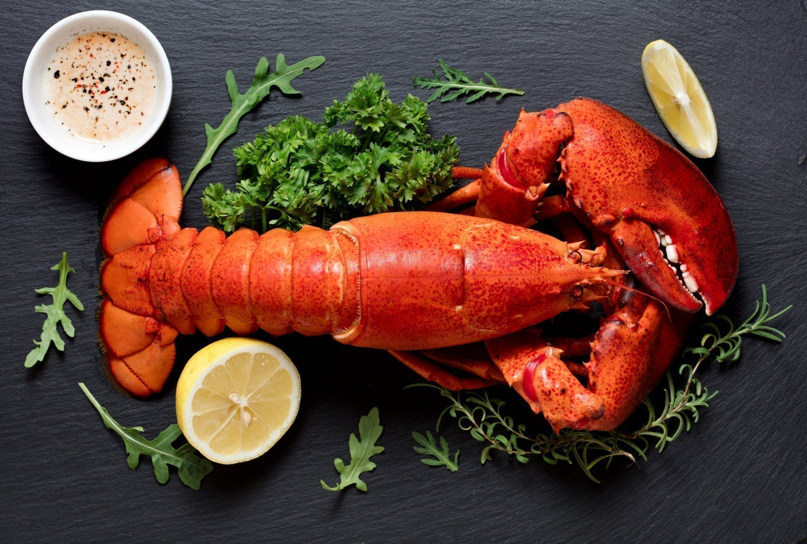 Cooked Maine lobster tail and claws served with lemon, dipping sauce, and fresh herbs on a black slate plate.