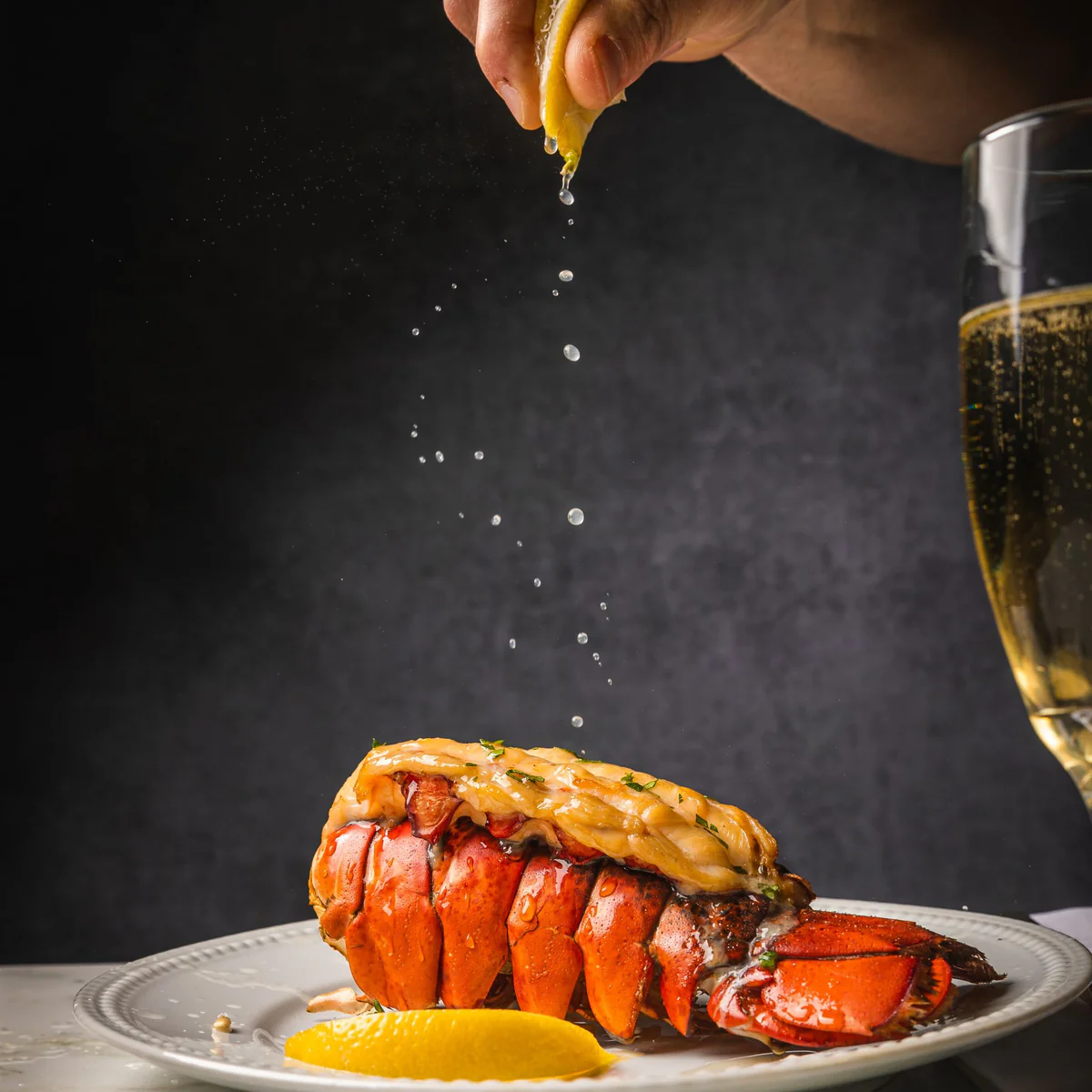 Sprinkling lemon on lobster tails, alongside a glass of champagne and a lemon wedge. 