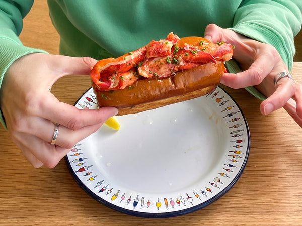 A person wearing a green sweater is holding a Chicago-style lobster roll while sitting near a wooden table.