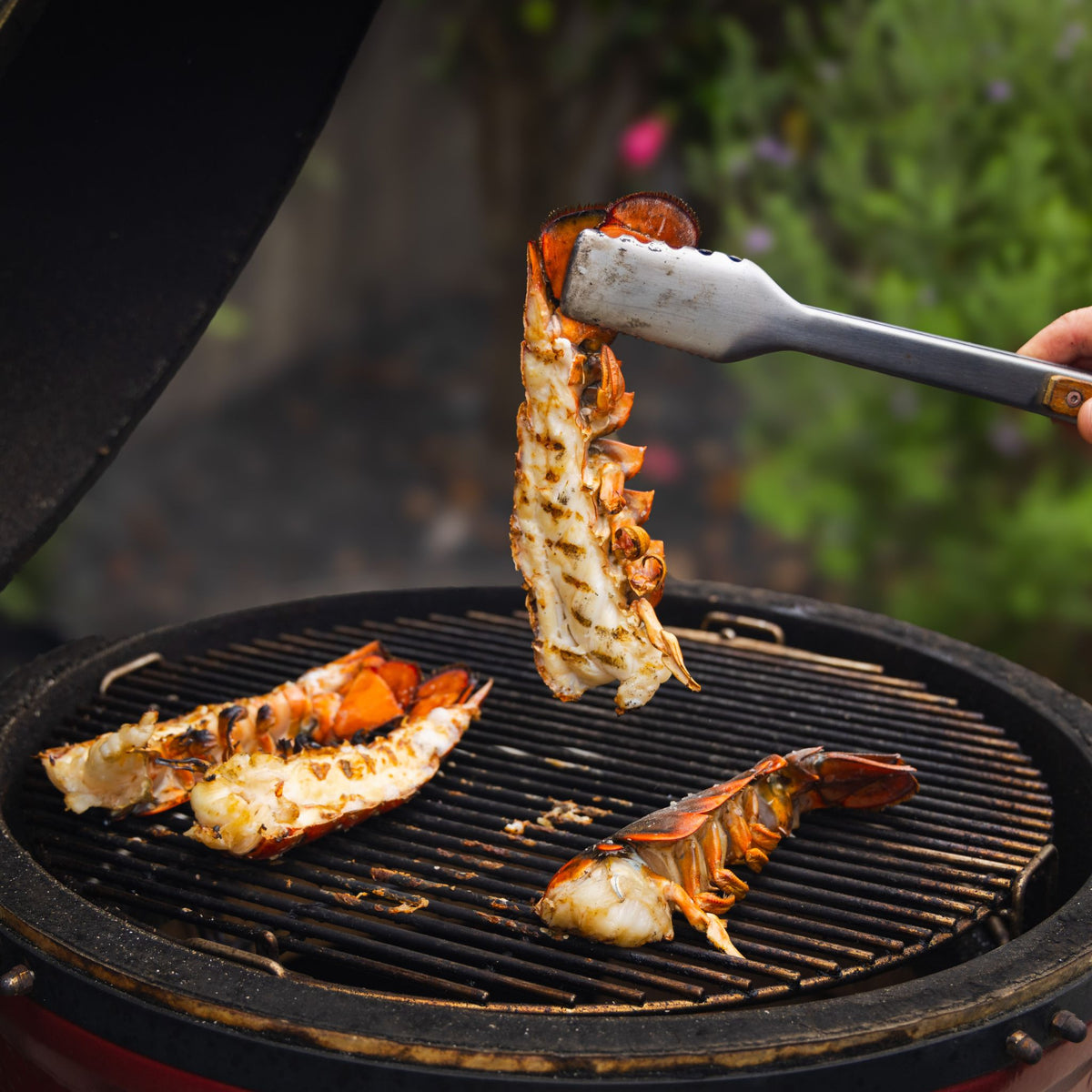 Lobster tails being flipped on a barbecue grill with tongs.